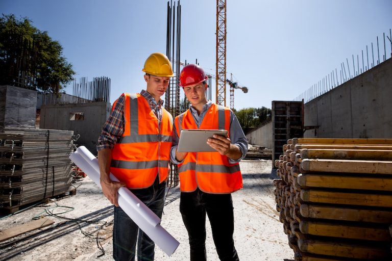 Civil architect  and construction manager  dressed  in orange work vests and  helmets discuss  a building project on the mobile tablet on the open building site next to the crane . .