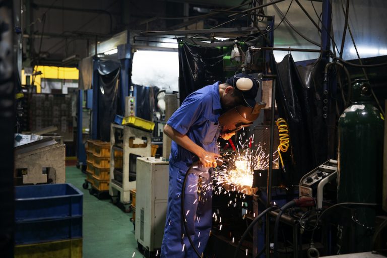 Man wearing blue overall and welding mask standing in factory, welding metal, sparks flying.